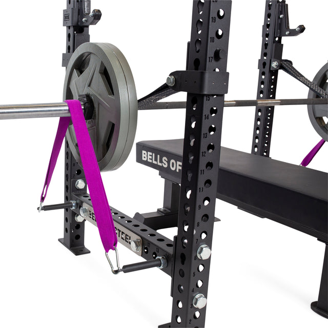 A close-up of a bench press setup featuring Bells of Steel Band Pegs with Carabiners, weight plates, a barbell, and a purple resistance band attached to the rack—ideal for enhanced resistance training.