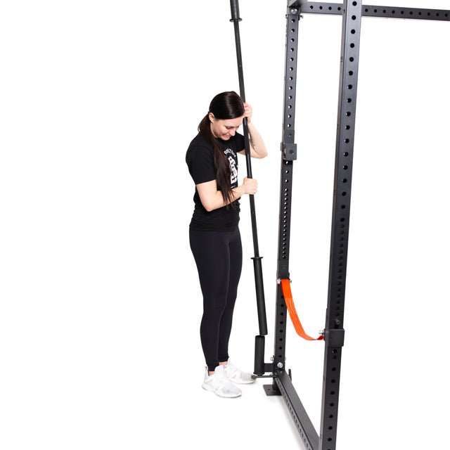 A woman in athletic wear adjusts a barbell on a Bells of Steel Vertical Mount Barbell Holder Rack Attachment, which is fixed to a black power rack with an orange safety arm, all on a white background.