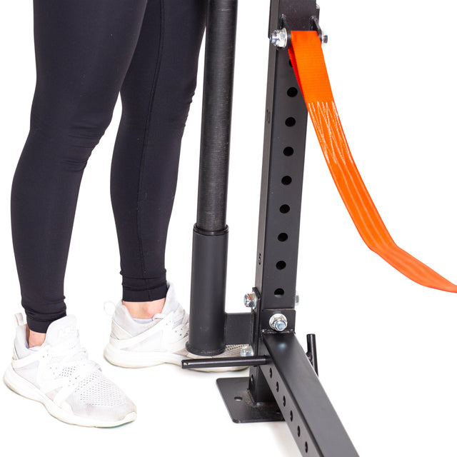 A person in black leggings and white sneakers stands next to the Bells of Steel Vertical Mount Barbell Holder Rack Attachment, shown attached to a black metal gym rack with an orange strap against a white background.