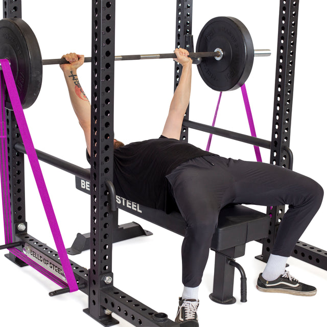 A person bench presses a barbell inside a power rack, using Bells of Steel Standard Band Pegs with resistance bands attached for support. Pink safety straps are in place. The lifter wears a black shirt, joggers, and sneakers.