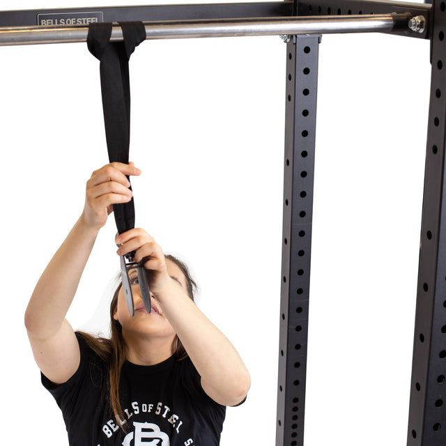 Wearing a black Bells of Steel shirt, a person loops the Cable Pulley’s black strap over a power rack’s pull-up bar, preparing for Lat Pulldowns or Tricep Pushdowns against a white background.