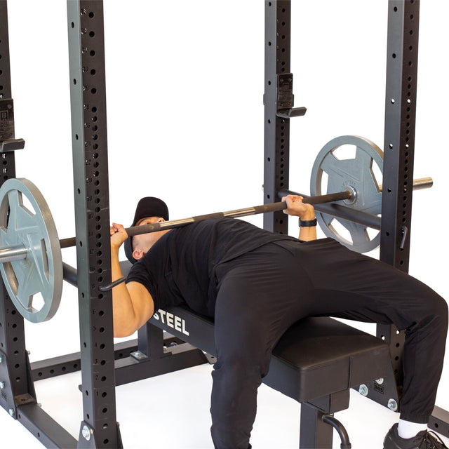 Wearing black workout gear, a person bench presses with a barbell and weight plates on the Bells of Steel Hydra Flat Foot Power Rack Builder (3" x 3", ⅝" holes), showcasing a key home gym movement.