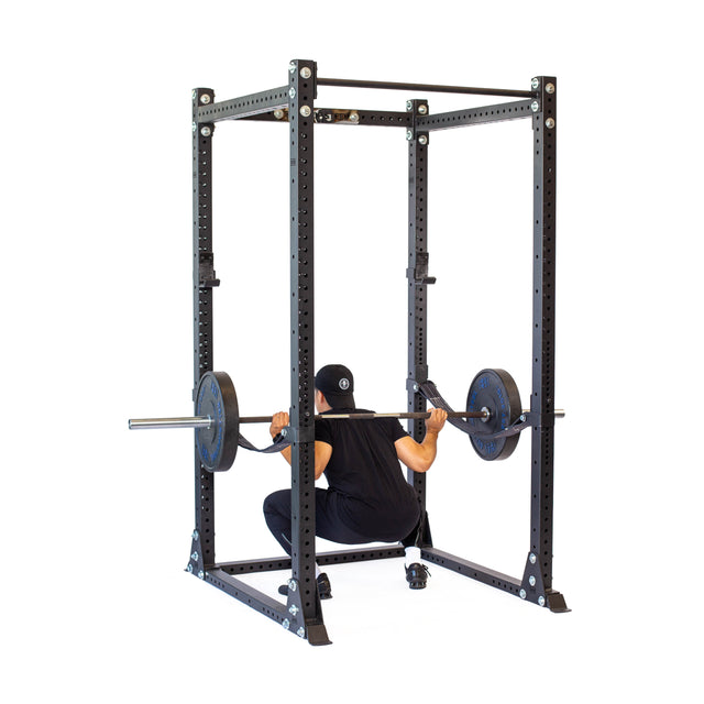 A person in black workout clothes and a cap performs a barbell squat inside the Bells of Steel Hydra Flat Foot Power Rack Builder (3" x 3", ⅝" Holes) with weight plates, ideal for a home gym.