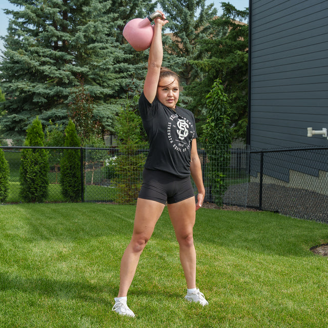 A woman stands on grass, holding a pink Bells of Steel Competition Kettlebell overhead with one arm. She wears a black t-shirt, black shorts, and white shoes. Trees and a fence are visible in the background.