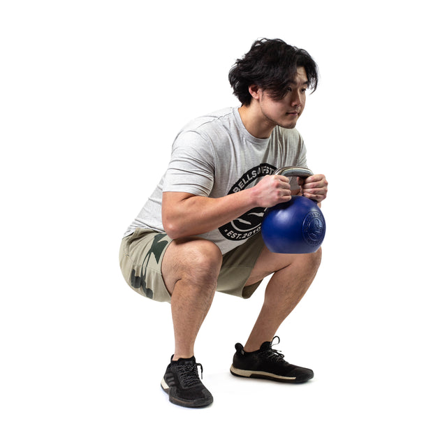 A man in a gray t-shirt, camo shorts, and black sneakers squats while gripping a blue Bells of Steel Competition Kettlebell with both hands against a white background, emphasizing his dedication to kettlebell sport training.