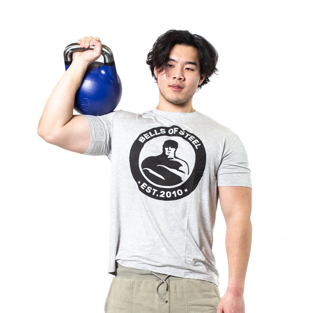 A person in a grey Bells of Steel t-shirt holds a blue Bells of Steel Competition Kettlebell on their shoulder, standing against a plain white background.