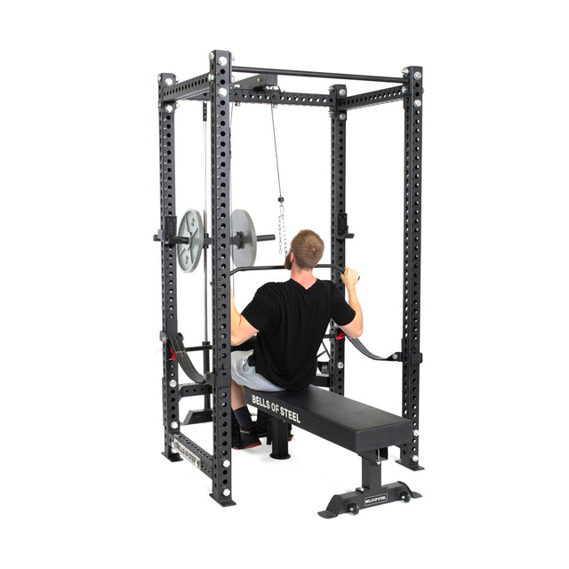 A person uses the Bells of Steel Lat Pulldown & Low Row Rack Attachment - Hydra & Manticore with Olympic weight plates on a power rack, performing cable exercises like lat pulldowns on a workout bench.