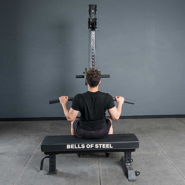 A person sits on a Bells of Steel bench, gripping the Fat Bar - Lat Pulldown Bar attached to a lat pulldown machine, ready to boost grip strength in a gym with gray walls and flooring.