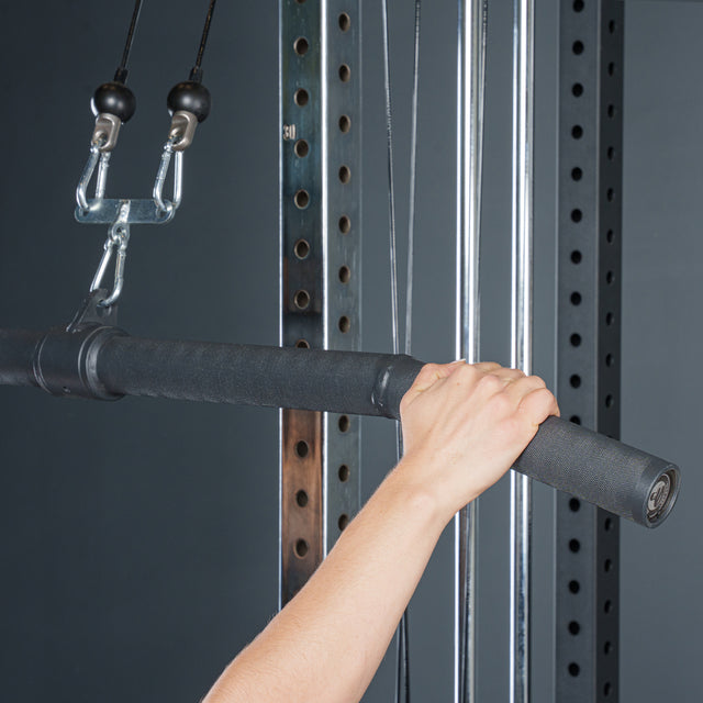 A close-up of a hand gripping the Bells of Steel Fat Bar - Lat Pulldown Bar, a thick black cable machine attachment that enhances grip strength, shown attached to cables and pulleys in a dark gym.