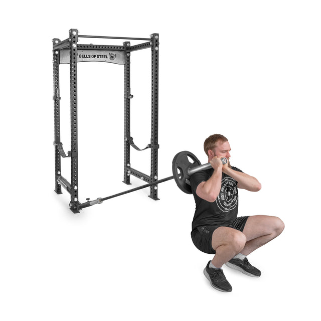 A person performs a front squat with a barbell and plates, displaying their strength routine in front of a Bells of Steel metal squat rack. They sport a black T-shirt, shorts, and sneakers against a white backdrop. The setup suggests the use of UHMW protection for equipment longevity.