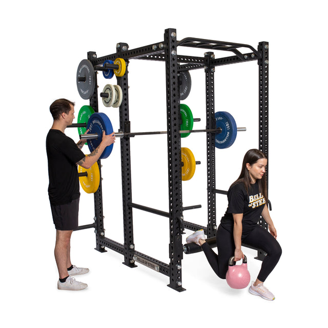 A man adjusts a barbell on the Manticore Six Post Power Rack by Bells of Steel, covered with vivid weight plates. Nearby, a woman in athletic gear gracefully lunges with a pink kettlebell, embodying the dynamic energy of their gym's modular setup for flexibility and strength.