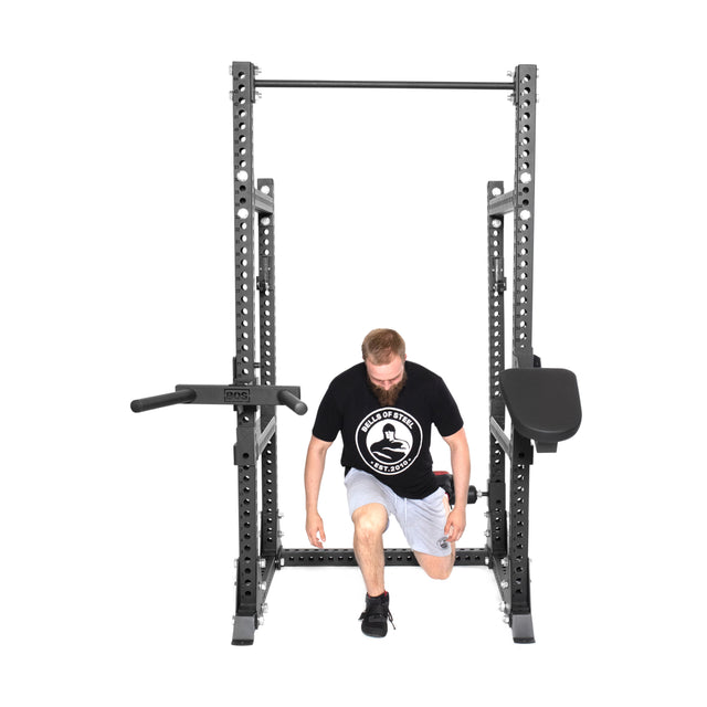 A man in a black t-shirt and white shorts kneels inside a Bells of Steel Manticore Half Rack - Prebuilt (3" x 3", 1" Holes) with multiple attachments, including bars and pads, on a white background.