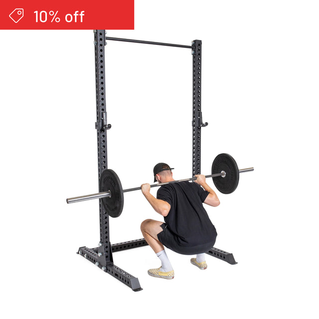 A person in a black shirt and cap squats inside the Bells of Steel Manticore Squat Stand Builder (3" x 3", 1" holes). A red “10% off” banner appears top left—perfect for your home gym.