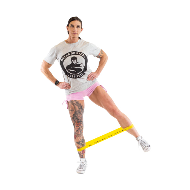 A woman demonstrates Bells of Steel Mini Bands by standing on one leg with the yellow band around her ankles, raising the other leg to the side. She sports a gray t-shirt, pink shorts, sneakers, and a tattoo on her left leg.