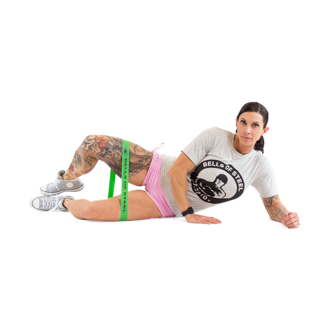 A tattooed woman in a gray T-shirt, pink shorts, and sneakers demonstrates side-lying clamshells with green Bells of Steel Mini Bands around her thighs against a white background, showcasing the versatile gym accessory.