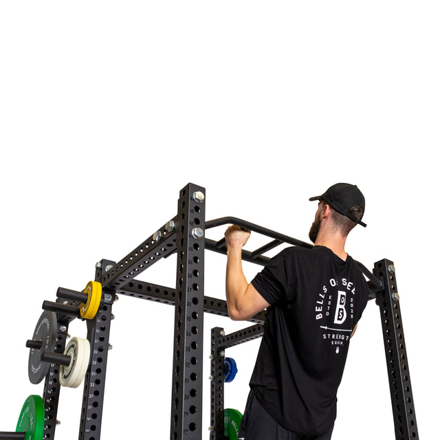 A man in a black T-shirt and cap performs a pull-up on the Bells of Steel Multi-Grip Pull Up Bar, which is mounted on a black power rack with weight plates, showcasing strength training against a white background.