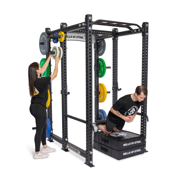 A man kneels on a padded platform inside the Manticore Six Post Power Rack Builder from Bells of Steel, exercising. A woman adjusts weight plates on the bar's modular components. Various colored plates are stacked on the rack pegs, with both wearing athletic clothing.