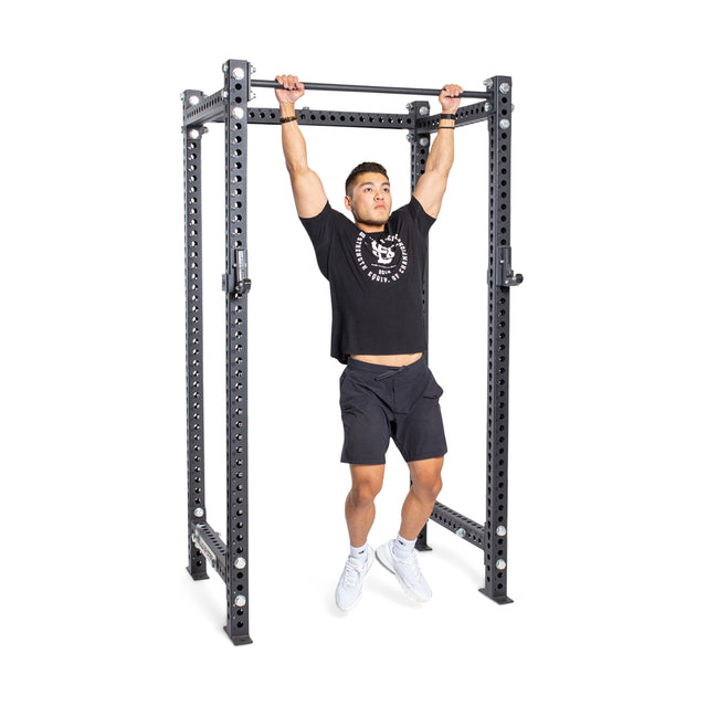 A person in a black T-shirt and shorts demonstrates their strength by doing a pull-up on the durable Straight Pull Up Bar by Bells of Steel. Mid-rep with arms extended and legs straight, they're wearing white athletic shoes against a plain white background.