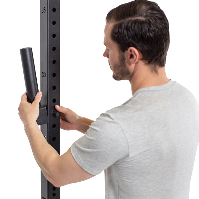 A man in a light gray shirt adjusts Bells of Steel Pin Plate Pegs on a vertical fitness rack, aligning them with numbered holes for organized Olympic weight plate storage.