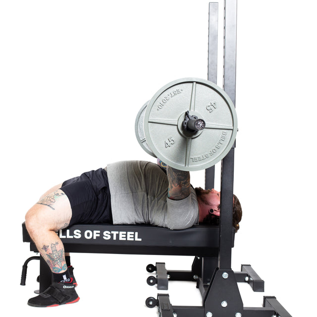A tattooed person bench presses on a Bells of Steel bench, lifting the Bells of Steel Powerlifting Bar with weight plates, viewed from the side against a white background.