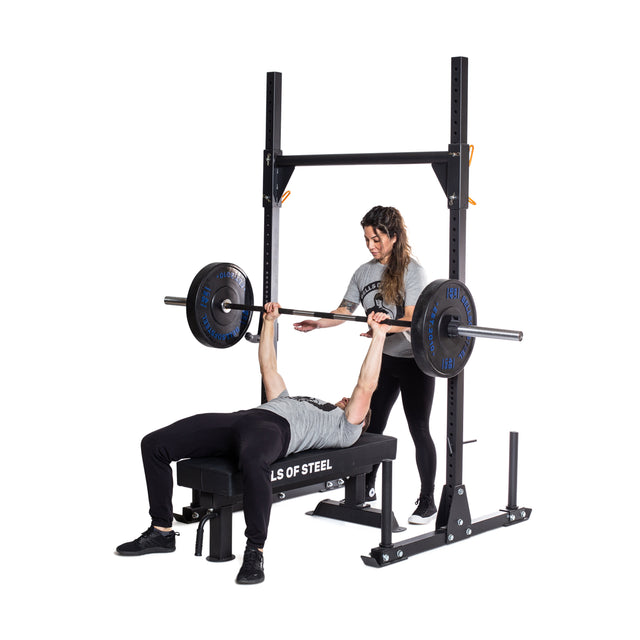 A person bench presses on a weight bench using the Bells of Steel Powerlifting Bar, while a spotter stands behind. Both wear athletic gear, and the Bells of Steel Powerlifting Bar is featured against a plain white background.