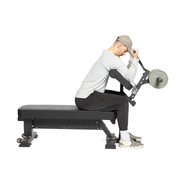A person in a gray shirt, black pants, and checkered shoes sits on Bells of Steel Bench Attachments performing a preacher curl with a barbell. The "BELLS OF STEEL" branding is visible against a plain white background.