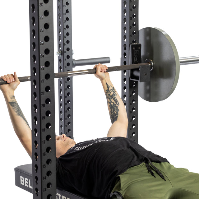 A person with tattooed arms lies on a bench, gripping a barbell held by Bells of Steel Roller J-Cups in a power rack. Wearing a black shirt and green pants, they benefit from safe lifting and barbell protection before starting their set.