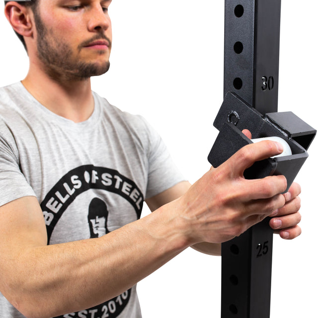 A man in a grey Bells of Steel t-shirt adjusts Roller J-Cups onto a squat rack with numbered holes, showcasing how these attachments protect the barbell during bench presses and other lifts.
