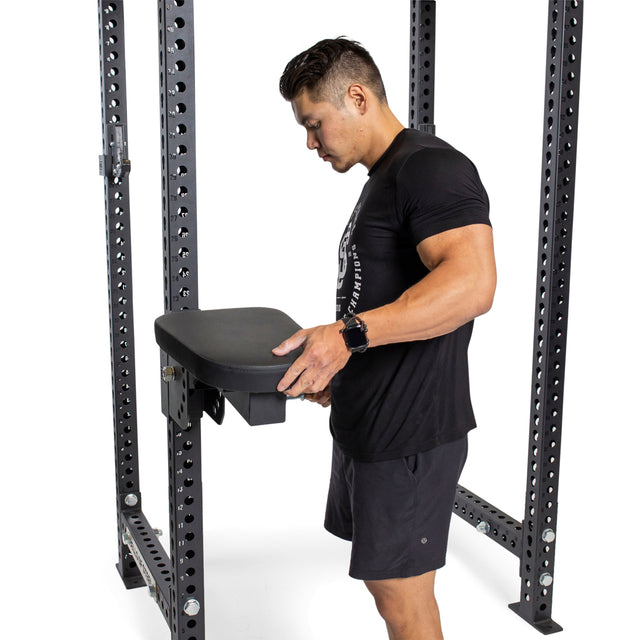A man in athletic wear adjusts the Bells of Steel Seal Row Pad Rack Attachment, a chest support added to a metal power rack, preparing the gym equipment for use.