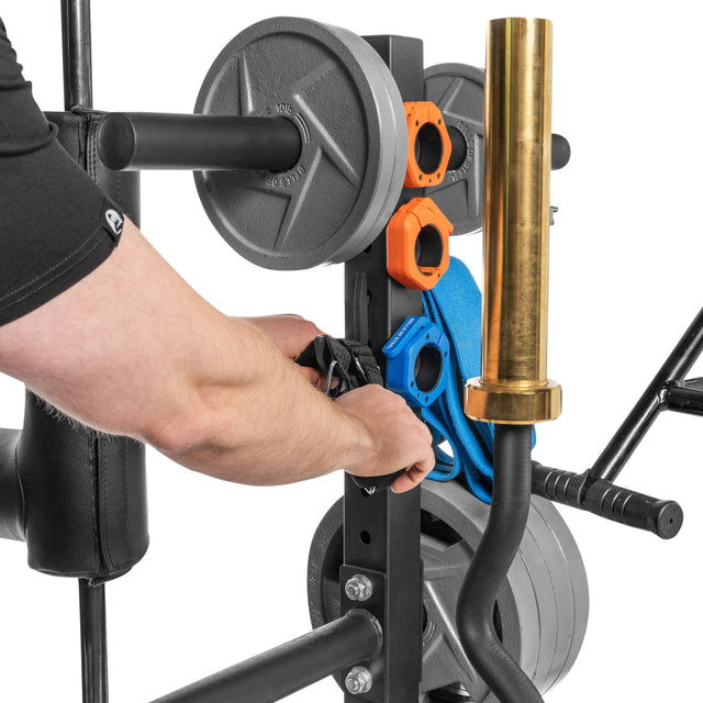 In their home gym, someone adjusts blue and orange safety gear on a metal apparatus loaded with weight plates and a gold barbell. The focus is on the hands and equipment neatly organized by the Bells of Steel Residential Bumper Plate Tree.