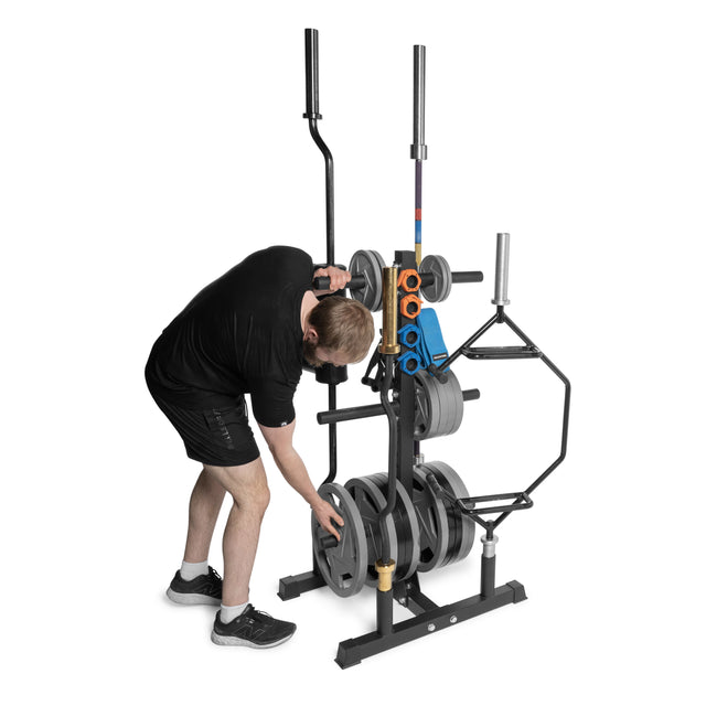 Dressed in black, a man organizes weights on his Bells of Steel Residential Bumper Plate Tree. The rack, designed for convenient weight plate storage, features multiple plates and handles. He is bent over adjusting them in a home gym setting.