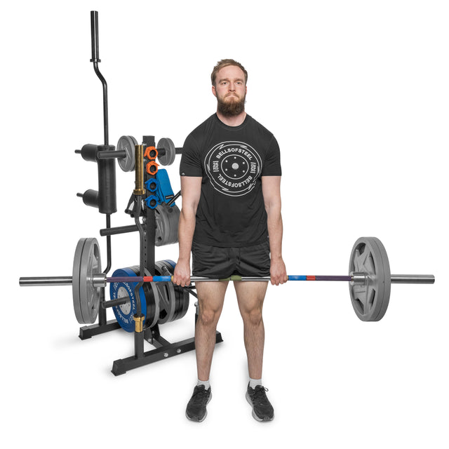 A man with a focused expression lifts a barbell on a platform in a gym. He's wearing black shorts and a shirt against the white background. Next to him is the Bells of Steel Residential Bumper Plate Tree for convenient plate storage alongside other home gym equipment.