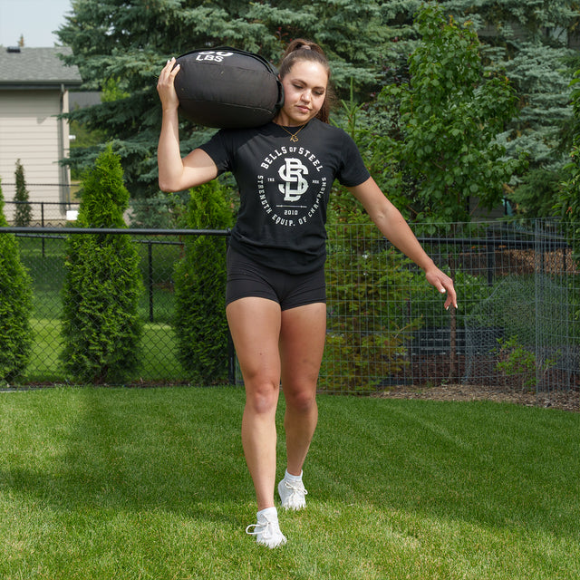 A woman makes the most of her outdoor workout with the Bells of Steel Ultimate Backyard Training Bundle, carrying a heavy sandbag on her shoulder while walking on grass in a backyard gym, dressed in a black t-shirt, black shorts, and white sneakers.