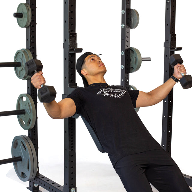 A man wearing a black t-shirt and cap uses the Bells of Steel Seal Row Pad Rack Attachment on a bench inside a power rack, with weight plates visible behind him.