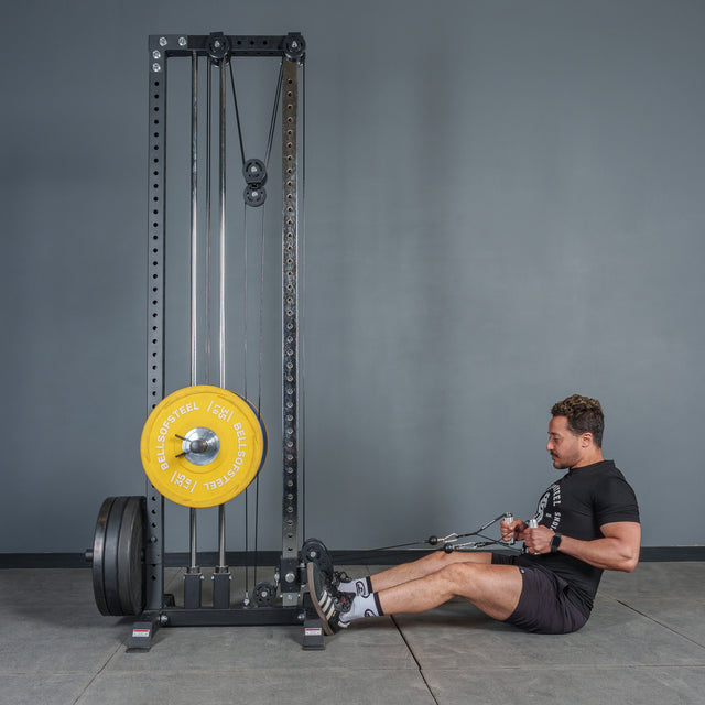 A man in a black shirt and shorts uses the Bells of Steel Aluminum Handles Pair with Bearings to perform a seated cable row on a weight machine with yellow and black plates, set against a plain gray wall.