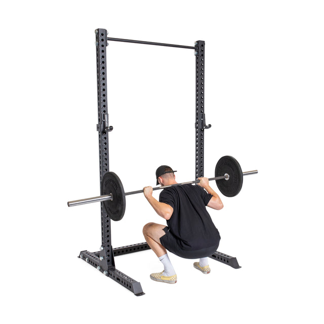 A person in black attire is squatting with a barbell inside the Bells of Steel Manticore Squat Stand Builder (3" x 3", 1" holes) against a plain white background.