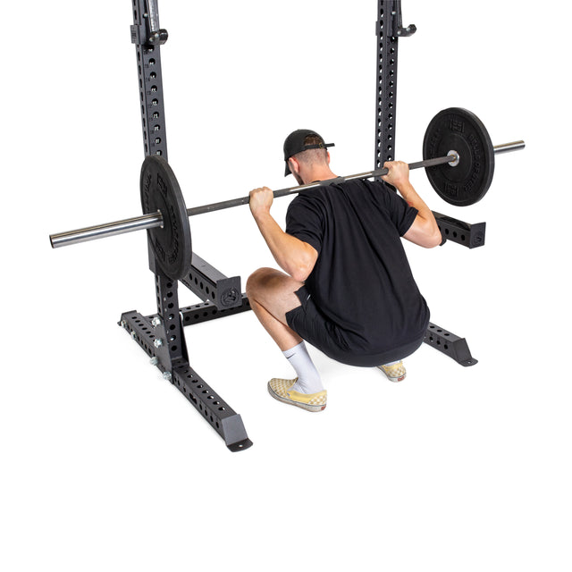 A man in black workout attire performs a deep squat with a barbell on his upper back using the Bells of Steel Manticore Squat Stand Builder (3" x 3", 1" Holes) inside his home gym.