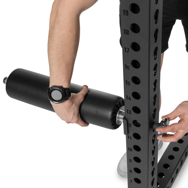 A person adjusts the Bells of Steel Split Squat Leg Roller Rack Attachment on a metal exercise rack with numbered holes, using their left hand to tighten a bolt while wearing a black watch, in preparation for Bulgarian split squats.