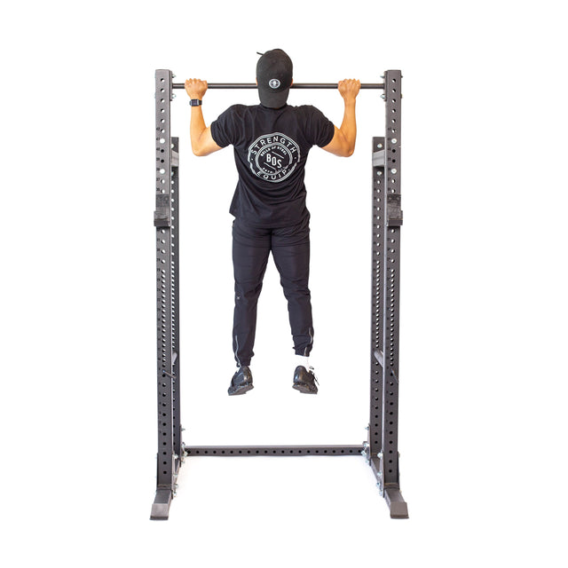 Clad in a black shirt with a circular logo, matching pants, and a cap, a person grips the Bells of Steel Straight Pull Up Bar. Their focus and form stand out against the white backdrop.