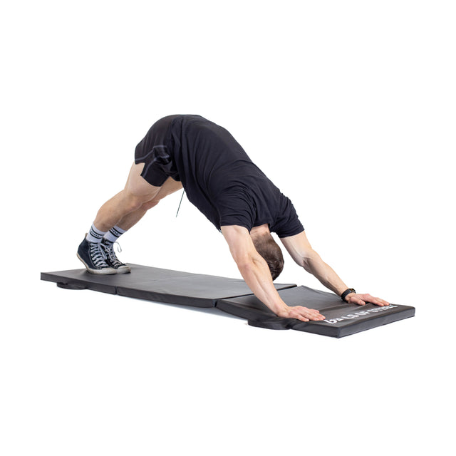 A person in black athletic wear and shoes performs a downward dog yoga pose on the Bells of Steel Trifold Mat against a white background.