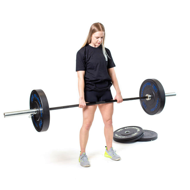 A person powerlifting the Bells of Steel Multi-Purpose Olympic Barbell with black weights in a bright studio. They're wearing a black shirt, shorts, and gray sneakers with yellow accents. Nearby, extra weight plates rest on the floor.