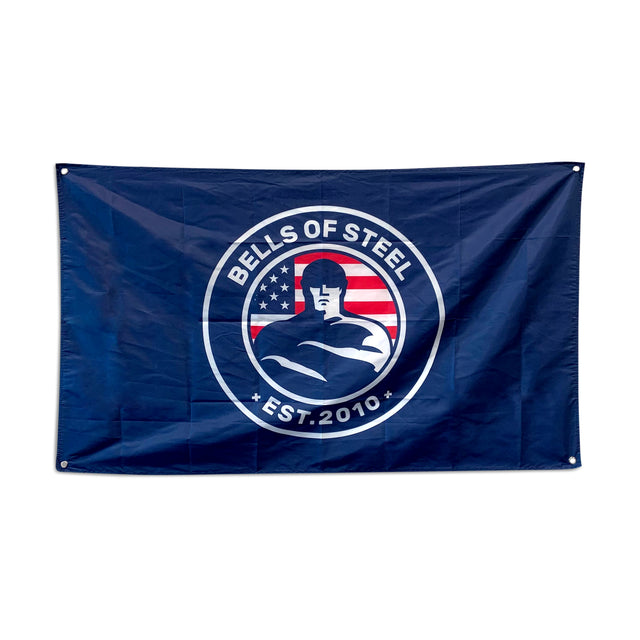 The Bells of Steel Flags feature a navy blue design with a circular logo of a muscular figure, an American flag backdrop, and "Bells of Steel Est. 2010"—an inspiring addition to any home gym.