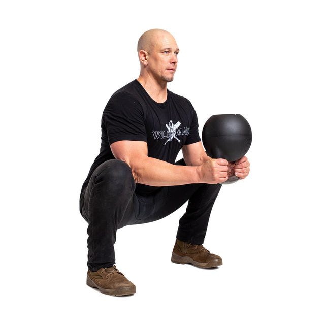 In a contrasting white setting, an individual in black attire performs a squat with intense focus using the Bells of Steel Mark Wildman Adjustable Kettlebell, showcasing their dedication to strength and precision.