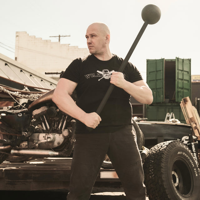 A muscular man showcases his core strength standing on a flatbed truck, gripping a Bells of Steel Adjustable Macebell. Wearing a black t-shirt and pants, he's framed by a motorcycle and an open shipping container, creating an industrial outdoor scene.
