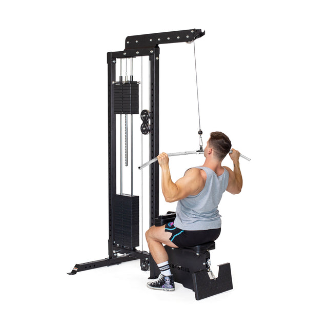 A man uses the Bells of Steel Lat Pulldown Bar on a cable machine, pulling it toward his chest to train his back. He wears a gray tank top, black shorts, and sneakers against a plain white background.