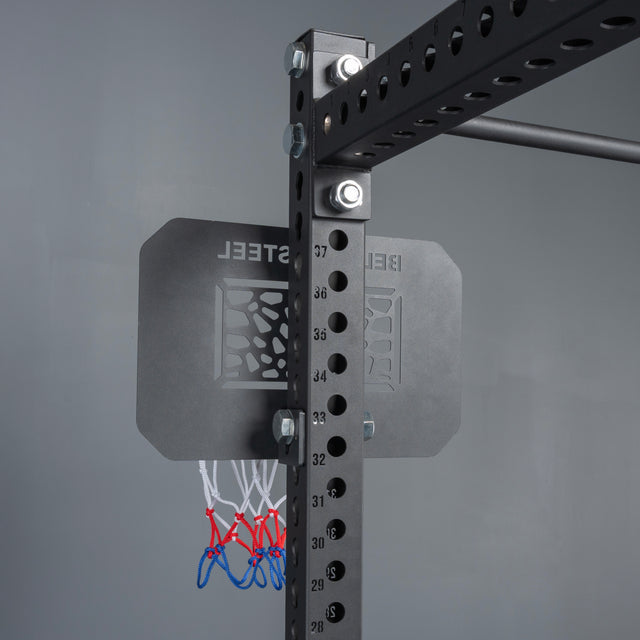 The Bells of Steel Rack Attached Basketball Hoop w/ Ball, featuring a small metal hoop, black backboard, red-white-blue net, and numbered holes, is mounted to a power rack against a gray wall.