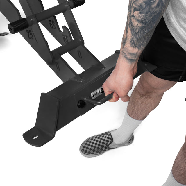 Wearing a gray shirt, black shorts, and checkered slip-ons, a tattooed individual grips a gym machine with 25 lb plates beside the Buzz-Saw Heavy-Duty Adjustable Bench from Bells of Steel, ready for an intense workout.