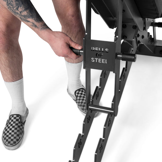 A person in white socks and checkered slip-on shoes adjusts the height of a Bells of Steel Buzz-Saw Heavy-Duty Adjustable Bench by moving a pin into numbered slots.