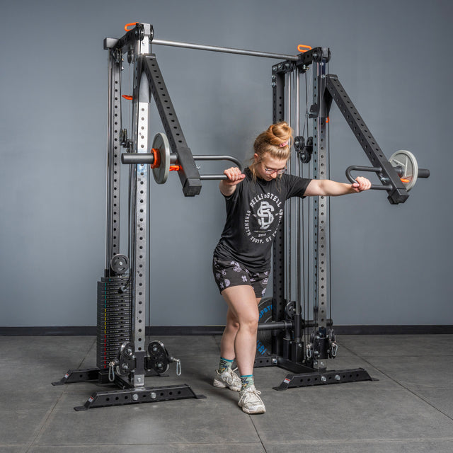 A woman performs a chest press in the gym using the Bells of Steel Lever Arms Rack Attachment, with weights attached. She is dressed in a black t-shirt, printed shorts, socks, and athletic shoes.
