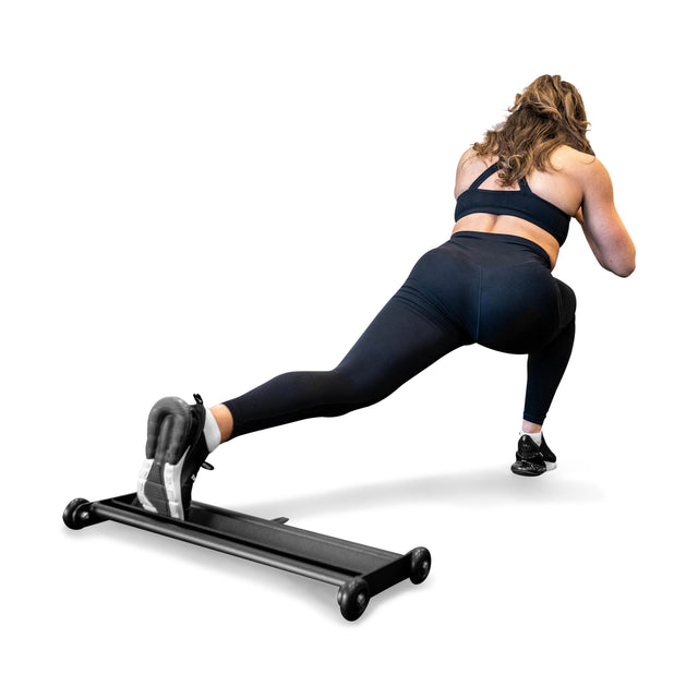 A woman in black workout clothes uses the Bells of Steel Glute Ham Slider to perform a reverse lunge, engaging her posterior chain, with one foot on the platform and the other leg bent, set against a white background.